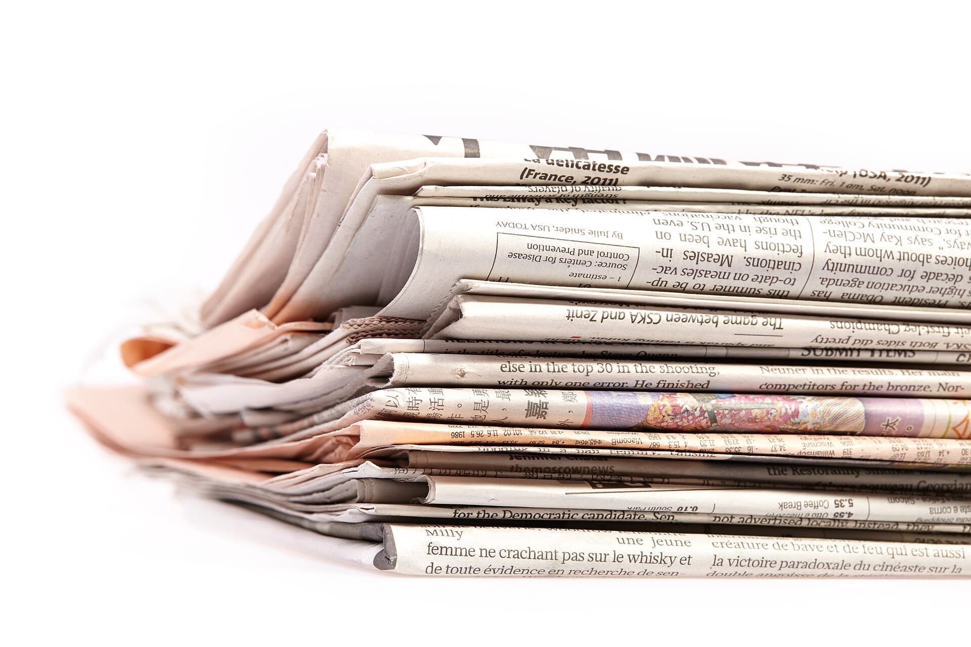 A stack of folded newspapers in several languages, photographed close up against a white background